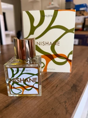 Nishane Papilefiko extrait de parfum 100ml - Nishane Papilefiko Extrait de parfum i en stilren, fyrkantig glasflaska med guldfärgad metallkork. Flaskan har en modern etikett med gröna och orange slingor och matchande förpackning. En exklusiv doft för dig som gillar unika parfymer.