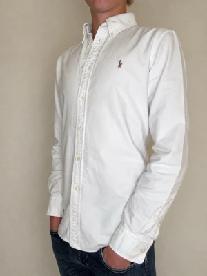 Ralph Lauren skjorta - Säljer en vit skjorta från märket Ralph Lauren. Skjortan är i fint skick, den är varsamt använd utan deffekter. Passformen är custom fit. Storleken är L men skjortan passar mindre storlekar, exempelvis M. Denna skjorta passar perfekt året om och nypris är ca 2000kr! Kontakta mig vid frågor eller prisförslag!