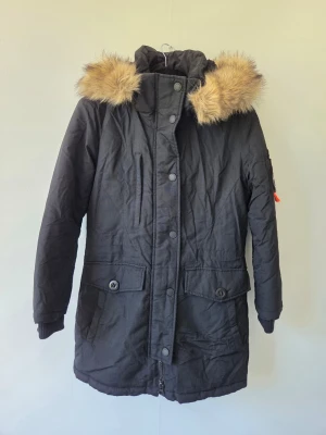 Super dry Svart parkas Everest - Svart parkas med huva och fuskpälsdetalj. Jackan har stora framfickor med knappar, dragkedja och tryckknappar framtill samt ribbade muddar vid ärmsluten. Perfekt för kalla dagar och har en klassisk, tidlös look. Litet hål i sista bilden.