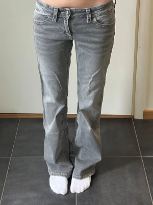 Grå bootcut jeans med låg midja - Säljer ett par grå jeans med bootcut-modell från Gina Young i storlek 164. på ena benet har de lite slitningar så de har släpat i marken, men på andra benet finns det inga slitningar. 