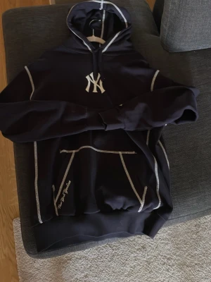 Mörkblå Yankees hoodie med tryck - Oanvänd mörkblå hoodie med New York Yankees-logga framtill och stort 'World Series 2009' tryck på ryggen. Hoodien har vita kontrastsömmar, magficka och huva med dragsko. Perfekt för dig som gillar sportig streetstyle och baseball.