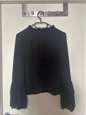 Svart blus med volangärmar - Stilren svart blus med hög hals och långa, vida ärmar med volangdetaljer. Blusen har en lätt transparent känsla och knäppning bak i nacken. Perfekt för dig som gillar en enkel men trendig look.