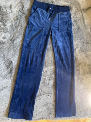 Marinblåa velourbyxor Juicy Couture - Säljer ett par marinblå mjukisbyxor från Juicy Couture i mjuk velour. Byxorna har bred resårmidja med dragsko, raka ben och stora fickor framtill. Perfekta för en chill och avslappnad stil.