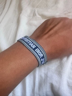 Christian Dior J'adior blått tygarmband - Snyggt blått tygarmband från Christian Dior med texten 'J'adior' broderad i vitt. Armbandet har justerbar knytning med tofsar i ändarna och en cool, avslappnad vibe. Perfekt accessoar för att lyfta din outfit med en touch av lyx.