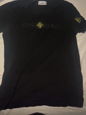 Svart Stone Island t-shirt - Snygg svart t-shirt från Stone Island med ikonisk logga broderad i gult och grönt på bröstet samt på ärmen. Klassisk passform och rund halsringning. Perfekt för dig som gillar streetwear och vill ha en stilren look.