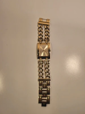 Guldig fyrkantig GUESS länkarmbandsklocka  - Stilren klocka med fyrkantig urtavla i guldton och dubbla kedjelänkar. Klockan har minimalistisk design utan siffror och ett metallarmband som ger en lyxig känsla. Perfekt accessoar för dig som gillar detaljer i guld.