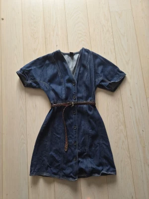 Jeansklänning från Lindex - Snygg jeansklänning från Lindex i mörkblå denim med kort ärm, v-ringning och knappar framtill. Det är storlek xs. OBS: skärp ingår EJ