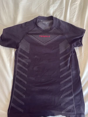 Svart tight tränings-tshirt YoungLA - Säljer en svart, tight tränings-tshirt från YoungLA med korta ärmar och diskreta gråa mönster längs sidorna. Materialet är stretchigt och andas, perfekt för gymmet eller löpning. Röd logga framtill ger en sportig touch. Säljer pga paus från gymmet 