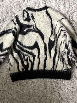 Stickad tröja med svartvitt mönster Zara - Säljer en fluffig stickad tröja från Zara i storlek S. Tröjan är vit med ett coolt abstrakt svart mönster och har ribbstickade muddar vid hals, ärmslut och nederkant. 