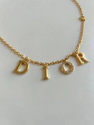 Guldhalsband med DIOR-bokstäver - Snyggt halsband i guldton med hängande bokstäver som stavar DIOR. O:et är dekorerat med små glittrande stenar för extra bling. Kedjan har även en liten stjärndetalj på sidan. Helt nytt.  Perfekt accessoar för dig som vill sticka ut.
