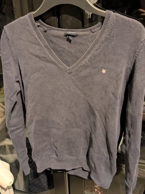 Svart stickad tröja från Gant - Mörkblå stickad tröja från Gant med V-ringning och diskret broderad logga på bröstet. Tröjan har långa ärmar och ribbade muddar vid ärmslut och nederkant. Lite urtvättad och har en liten fläck som säkert går att få bort i tvätten 