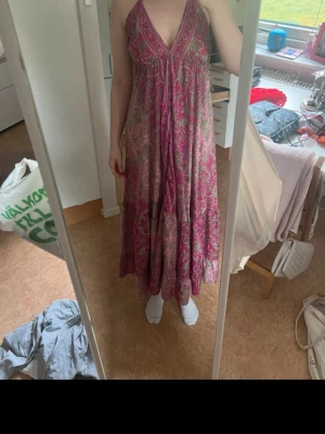 Rosa långklänning med snörning - Säljer en bohemisk långklänning i rosa och gröna toner med blommigt mönster. Klänningen har djup v-ringning, öppen rygg och snörning i nacken. Perfekt för sommardagar och festivaler. Flödande passform och tunnt, svalt material.