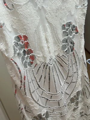 Vit paljettklänning med blommönster - Snygg vit kortklänning med glittriga paljetter i silver och inslag av rött. Klänningen har ett blommigt mönster och v-ringning. Perfekt för dig som vill sticka ut med en unik och festlig look. Materialet är troligtvis polyester med paljettbroderi.