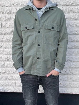 Olivgrön overshirt med fickor - Snygg olivgrön overshirt med raka linjer och två stora bröstfickor med lock och knappar. Klassisk krage och svarta knappar framtill. Tillverkad i ett slitstarkt bomullstyg som ger en avslappnad look. Perfekt att bära över en hoodie för extra lager. Storlek 44/S
