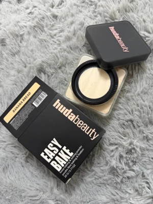 Huda Beauty Easy Bake Banana Bread - Lös puder från Huda Beauty i nyansen Banana Bread. Kommer i en fyrkantig plastburk med svart lock och rosa logga. Pudret har en ljusgul ton och är perfekt för att fixera foundation och ge en matt finish.