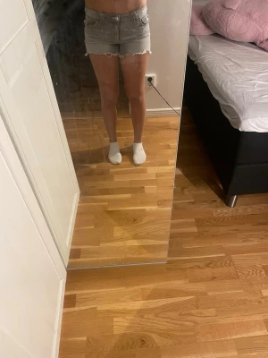 Grå jeansshorts med fransig kant - Säljer ett par ljusgrå jeansshorts med fransig, rå kant nertill och klassisk femficksdesign. Shortsen har normal passform och midja, samt detaljerade sömmar och knappar framtill. Perfekta för varma dagar och enkel att matcha med olika toppar.