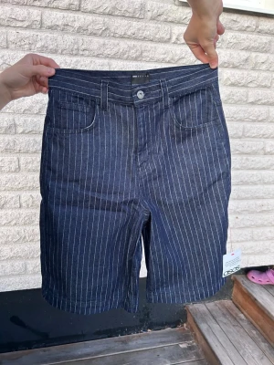 Mörkblå randiga Jeans-shorts  - Snygga mörkblå shorts med vita vertikala ränder och klassisk femficksdesign. Shortsen har normal passform, bälteshällor och stängs med knapp och dragkedja. Perfekta för sommaren och ger en clean och stilren look. Storlek EU 44