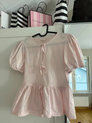 Ljusrosa topp med puffärm  - Helt oanvänd supersöt ljusrosa topp från Gina Tricot med puffärmar och volang nedtill. Toppen har tre knytband framtill och är gjord i mjukt bomullstyg. Perfekt för dig som gillar en trendig stil.