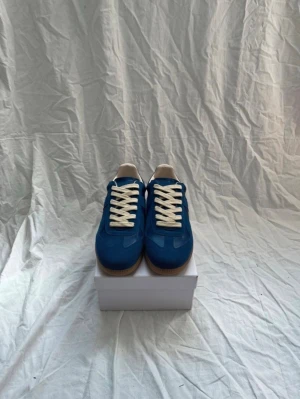 Blå gats - Snygga blå sneakers från Maison Martin Margiela med vita skosnören och beige detaljer vid hälen. Skorna har en klassisk låg siluett och gummisula i ljusbrunt med unikt mönster undertill. Perfekta för dig som vill ha något stilrent men ändå sticker ut.