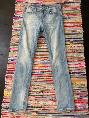 Ljusblå slim fit nudie jeans - Säljer ett par ljusblå slim fit nudie jeans med klassisk femficksdesign och snygga slitningar framtill. Jeansen har raka ben och subtila detaljer på bakfickorna. Perfekta för en avslappnad och trendig look.