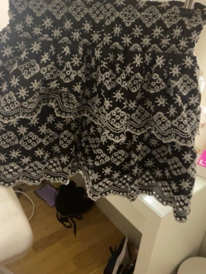 Svart kortkjol med vit brodyr - Kort svart kjol med detaljerad vit brodyr i blommigt mönster. Kjolen har en vågig kant nertill och elastisk midja för skön passform. Perfekt för dig som gillar unika detaljer och en feminin stil.