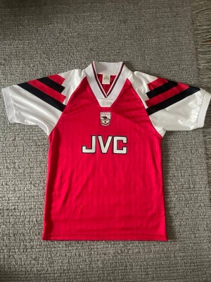 Arsenal retro matchtröja röd/vit - Klassisk Arsenal retro matchtröja i rött med vita ärmar och svarta samt röda detaljer. Tröjan har V-ringad krage med röd och vit kant, Arsenal-logga på bröstet och JVC-tryck framtill. Tillverkad i glansigt, lätt syntetmaterial. Perfekt för fotbollsfans!