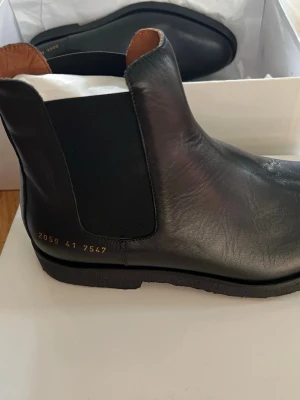 Svarta Chelsea boots från Common Projects - Stilrena svarta Chelsea boots från Common Projects i slätt läder med elastiska sidopaneler och diskret guldfärgad numrering på hälen. Skorna har en klassisk siluett, låg klack och svart yttersula. Levereras med originalkartong och dustbag.