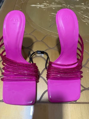 Rosa strasspumps med smala remmar - Säljer ett par trendiga rosa pumps från Zara med fyrkantig tå och smala remmar dekorerade med rosa strass. Skorna har hög klack och öppen häl, vilket ger en modern och snygg look. Perfekta för dig som vill sticka ut med färg och detaljer. Helt nya.