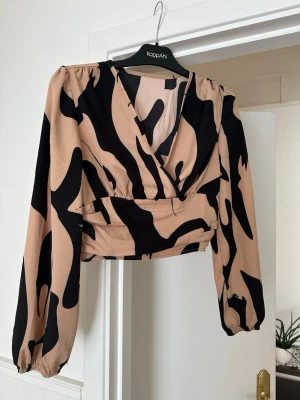 Beige och svart omlottopp, lite croppad - Trendig croppad omlottopp i beige och svart med zebrainspirerat mönster. Toppen har långa puffärmar, v-ringning och smock i ryggen för skön passform. Perfekt för dig som vill sticka ut med en unik look. Väldigt smickrande skuren, köpte för liten storlek, upplevs som en S (såldes som M). Lite stretchig och med sydda gummibandskanaler baktill, se bild 2. 