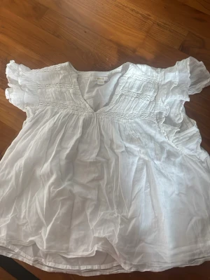 Vit blus med volangärm - Luftig vit blus i tunn bomull med v-ringning och fina volangärmar. Smockdetaljer upptill och lätt rynkad nedtill ger en romantisk och avslappnad look. Perfekt till jeans eller kjol för en trendig stil.