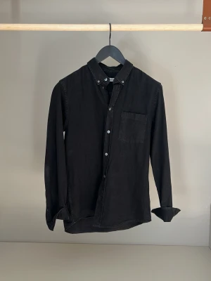 Svart långärmad skjorta i bomull från Wood Wood - Stilren svart skjorta från Wood Wood med klassisk krage och knappar framtill. Skjortan har en bröstficka och är tillverkad i mjuk bomull som . Perfekt till dig som gillar en enkel men snygg look. Långärmad modell med knapp vid ärmslut. 