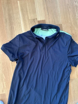 Mörkblå pikétröja med limegrön detalj - Snygg mörkblå pikétröja med krage och knappar framtill. Baksidan har en unik limegrön och vit panel upptill som ger en sportig look. Tillverkad i ett lätt och stretchigt syntetmaterial, perfekt för en avslappnad stil.