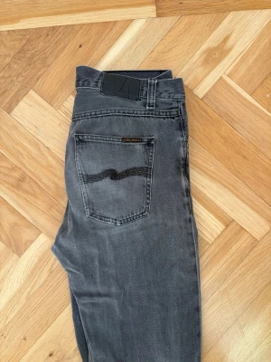 Grå jeans från Nudie Jeans - Snygga grå jeans från Nudie Jeans med klassisk femficksdesign och svarta detaljer på bakfickan. Jeansen har en smal passform och är tillverkade i denim med lätt tvättad look. Perfekta för en avslappnad och trendig stil.