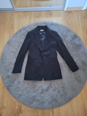 Svart kavaj från H&M, storlek 36 - Stilren svart kavaj från H&M med klassisk passform och smala slag. Kavajen har två fickor framtill och är helfodrad med slät insida. Perfekt för en clean och snygg look till vardags eller mer uppklätt.