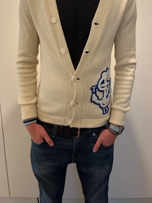 Beige kofta med blått broderi - Säljer en beige stickad kofta med djup V-ringning och knappar framtill. Koftan har ett stort blått broderat märke på vänster sida och ribbade muddar vid ärmslut och nederkant. Perfekt för dig som gillar en clean och stilren look.