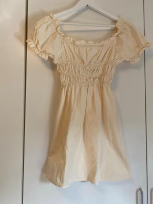 Ljus kort klänning från NA-KD - Supersöt ljus kort klänning från NA-KD med puffärmar och smock i midjan. Klänningen har volangdetaljer vid ärmslut och halsringning, och är tillverkad i ett mjukt bomullstyg. 