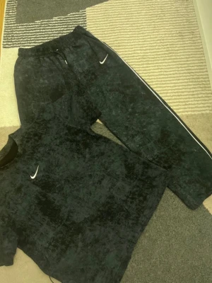 Svart Nike loungewear set - Säljer ett svart loungewear set från Nike med diskret mönster. Setet består av en t-shirt och ett par byxor med resår i midjan och vita detaljer längs sidorna. Både tröjan och byxorna har Nike-logga på bröstet respektive benet. Perfekt för chill dagar hemma. Storlek M 