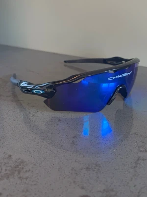 Oakley blå sportiga solglasögon - Säljer ett par Oakley solglasögon med blå spegelglas och svart båge. Modellen har en sportig, svepande form och tydlig Oakley-logga på sidan. Perfekta för dig som vill ha en cool och funktionell look till sommaren.