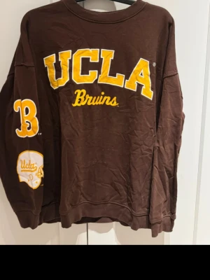 Brun UCLA Bruins hoodie från H&M - Säljer en brun hoodie från H&M Divided med stort gult UCLA Bruins-tryck på bröstet och siffran 3 samt hjälm på ärmen. Hoodien har ribbade muddar och är gjord i mjukt bomullsmaterial. Perfekt för dig som gillar college-stil och sportiga vibes.