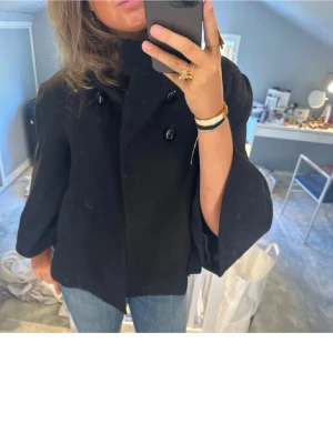 Svart trekvartsärmad kappa - Stilren svart kappa med dubbelknäppning och vida, trekvartsärmar. Kappan har en hög krage och en rak, lös passform som ger en cool och trendig siluett. Perfekt för dig som vill ha en unik och snygg kappa till hösten eller våren. Sparsamt använd. 