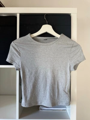Ljusgrå basic t-shirt - En enkel och stilren ljusgrå t-shirt. Modellen har korta ärmar och slutar över naveln. Använts ca 2 gånger. Finns tyvärr ingen lapp kvar i men är säker på att det är xs 