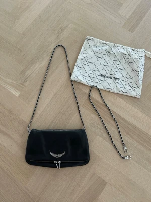 Svart axelväska Zadig & Voltaire - Snygg svart axelväska från Zadig & Voltaire i mjukt läder. Väskan har silverfärgade detaljer, kedjerem och ett coolt vingemblem framtill. Baksidan har en praktisk dragkedjeficka. Perfekt storlek för det viktigaste och levereras med dustbag.