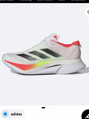 Adidas boston 12 - Vita adidas sneakers med färgdetaljer - Snygga vita sneakers från adidas med svarta ränder på sidan, röda och neongula detaljer samt en sportig, luftig mesh-överdel. Skon har en låg profil och en tjock, bekväm sula som ger bra stöd. Perfekt för dig som gillar en modern och sportig look. Storlek 39-40