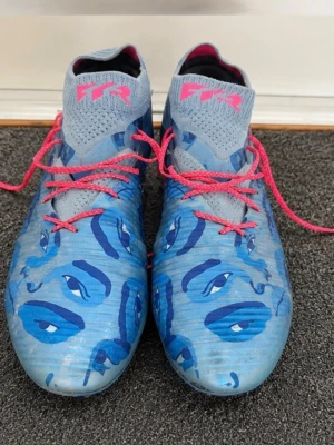 Puma fotbollskor - Säljer ett par snygga blåa fotbollsskor med rosa snören och detaljer. Skorna har en strumpliknande passform och mönstrad ovandel för extra grepp. Perfekta för dig som vill sticka ut på planen och ha bra kontroll på bollen.