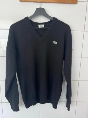 Svart V-ringad tröja från Lacoste - Klassisk svart V-ringad tröja från Lacoste med den ikoniska gröna krokodilloggan på bröstet. Tröjan har långa ärmar och ribbade muddar vid ärmslut och nederkant. Perfekt för en stilren och enkel look.