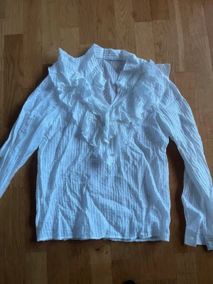 Vit blus med volangkrage - Säljer en vit blus med långa ärmar och en stor volangkrage framtill. Blusen har en lätt skrynklig struktur och v-ringning. Perfekt för dig som gillar romantiska detaljer och vill sticka ut lite extra.