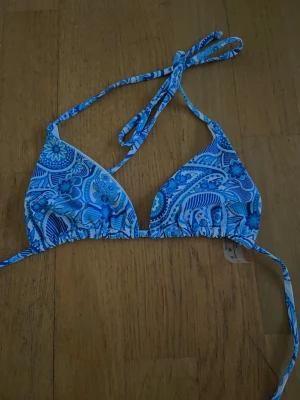 Blå paisleymönstrad bikiniöverdel XS - Snygg blå bikiniöverdel med paisleyinspirerat mönster i olika blå nyanser. Klassisk trekantsmodell med knyt i nacke och rygg. Tillverkad i mjuk polyester och elastan för skön passform. Perfekt för lata dagar på stranden eller poolen.