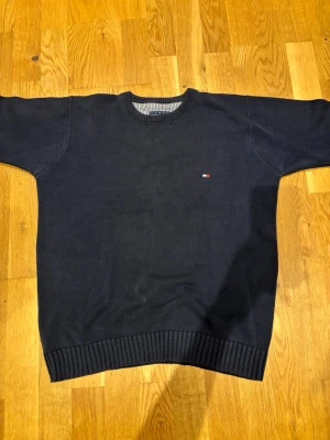 Mörkblå stickad tröja Tommy Hilfiger - Mörkblå stickad tröja från Tommy Hilfiger med rund halsringning och diskret logga broderad på bröstet. Tröjan har ribbade muddar vid ärmslut och nederkant. Perfekt för dig som gillar klassisk och stilren design.