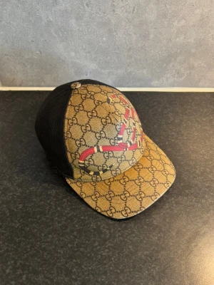 Gucci GG Supreme keps med orm  - Gucci-keps i klassiskt GG Supreme-mönster med orm-motiv framtill och svart mesh baktill.  Storlek XS / 56 cm. Justerbar baktill med Guccis gröna/röda web-detalj.  Säljes då den inte kommer till användning. Begagnat skick enligt bilder.  Observera: Kvitto, originalförpackning och övriga tillbehör finns inte kvar. Se samtliga bilder för skick och detaljer.