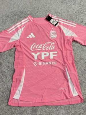Rosa Adidas fotbollströja Argentina - Säljer en rosa fotbollströja från Adidas med vita detaljer och tryck från Argentina landslaget. Tröjan har korta ärmar, rund hals och loggor från Coca-Cola, YPF och Binance. Tillverkad i lätt och ventilerande material, perfekt för träning eller match.